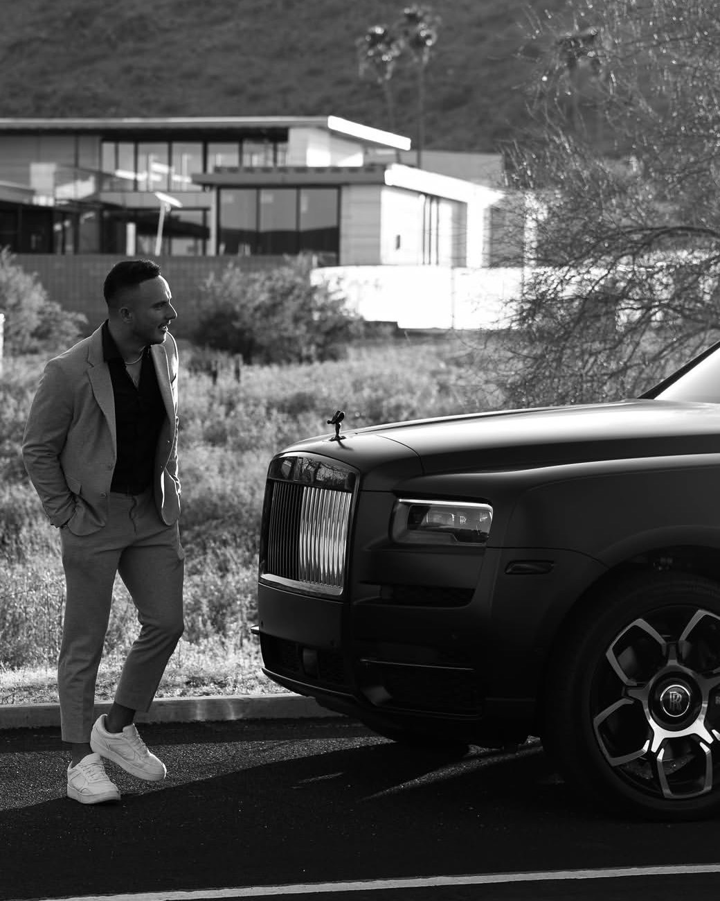 Jay Maska in black and white, standing beside a matte black Rolls-Royce Cullinan